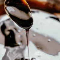 A silver spoon is coated in a thick, dark glaze. It is dripping back into a bowl that contains more dark glaze. The liquid in the bowl has a glossy sheen, reflecting the ambient light. The background is a soft blur, drawing the viewer's focus to the dripping sauce and its rich texture.