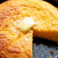 Cornbread in a cast iron pan with a triangle slice missing and melting butter cube in top in the centre.