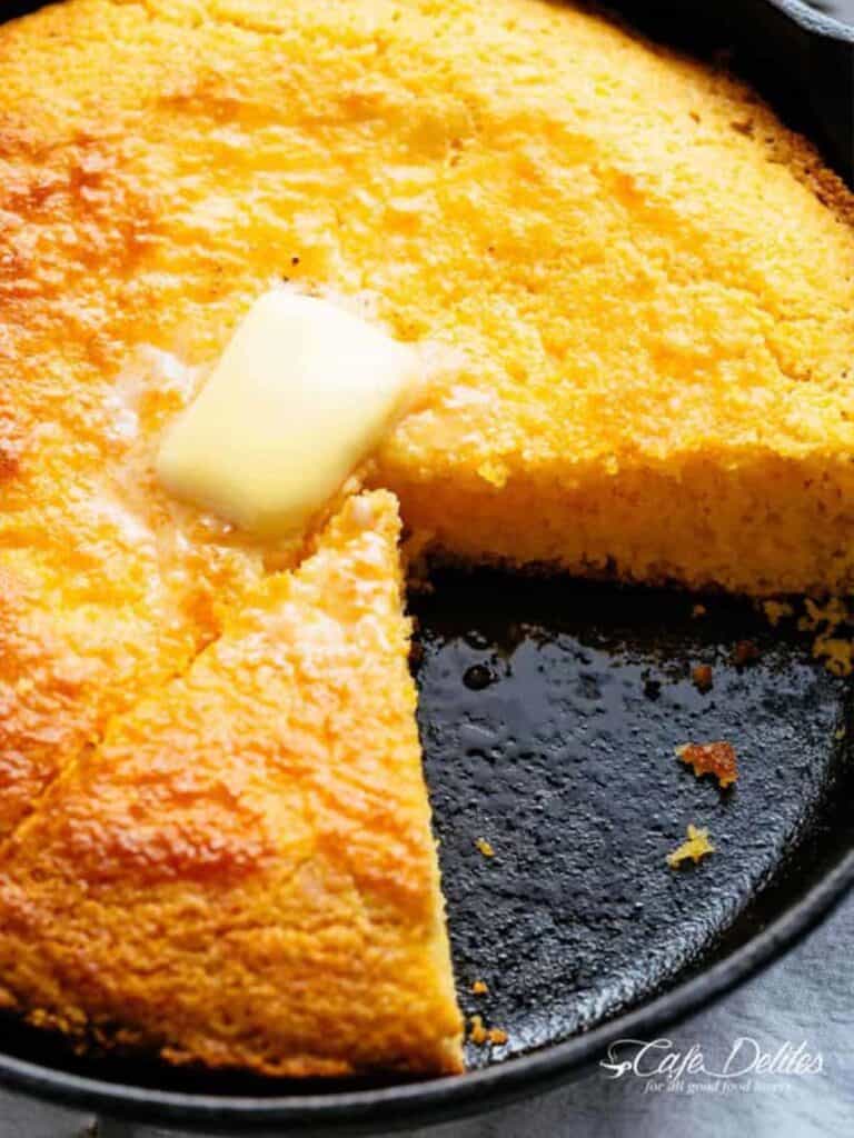 Moist cornbread in a skillet with butter piece in the centre and a slice cut out