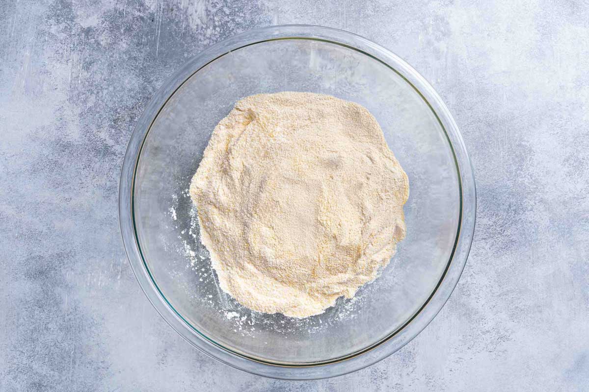 A picture shows polenta, flour, baking powder, baking soda, salt, and sugar mixed in a clear mixing bowl.