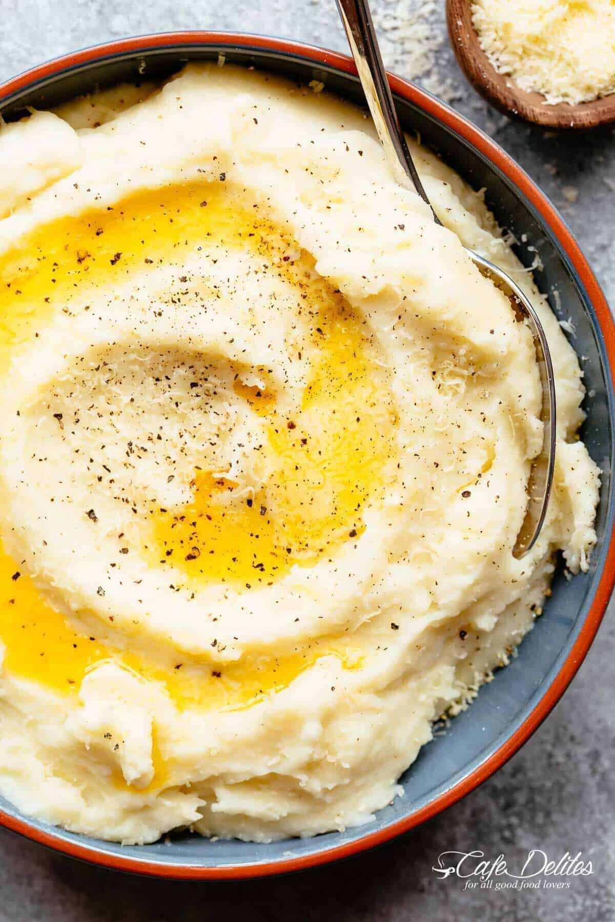 Keto Mashed Cauliflower with Garlic, Sour Cream and Parmesan #cafedelites #cauliflower #Keto #mashedcauliflower #mashedpotatoes Image of Mashed Cauliflower served in a grey bowl, topped with cracked pepper, oil, and Parmesan cheese, being scooped up by a silver spoon.
