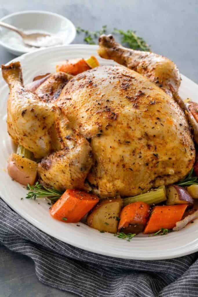 A full size slow cooked, golden chicken on a bed of roughly chopped carrots, potatoes and herbs, on a white serving platter.