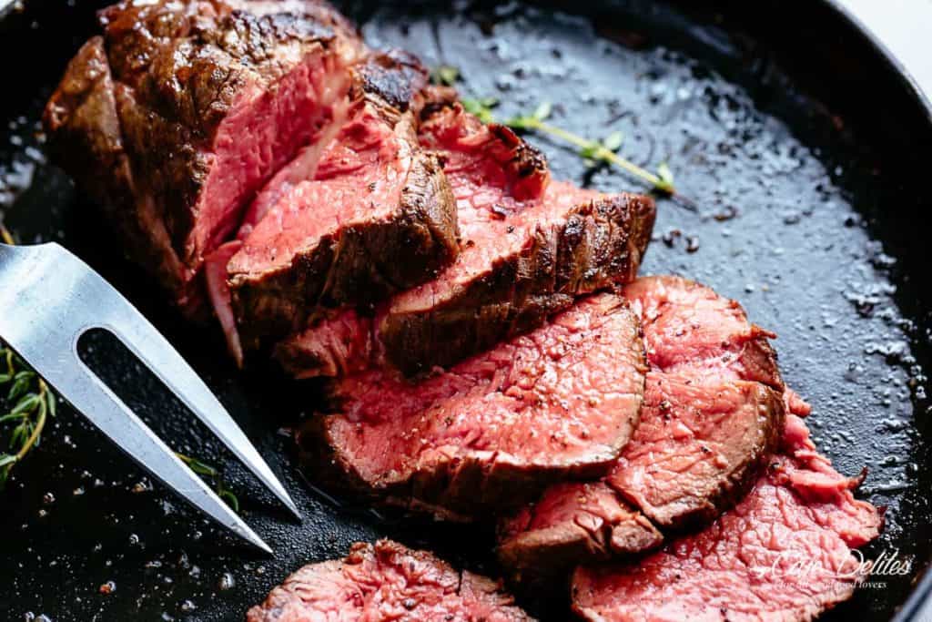 Beef Roast in a black skillet cut into slices cooked to medium rare with sprigs of fresh rosemary and a crack of pepper over the meat.