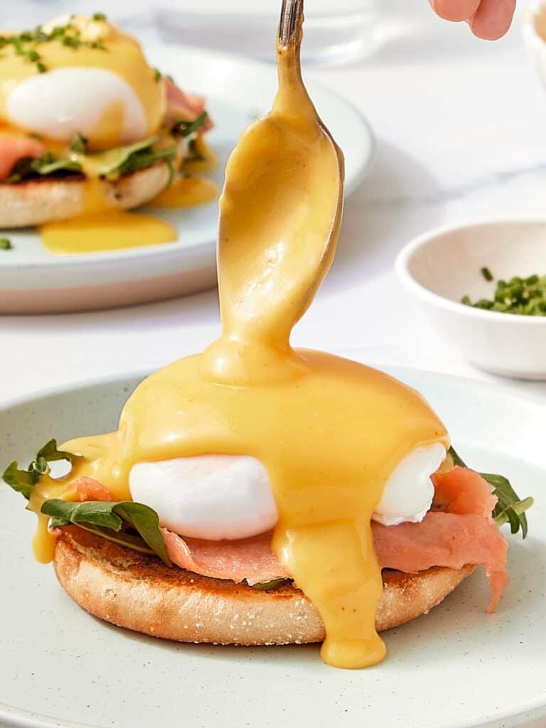 Golden Hollandaise sauce being spooned over a poached egg on an English muffin with smoked salmon and arugula.