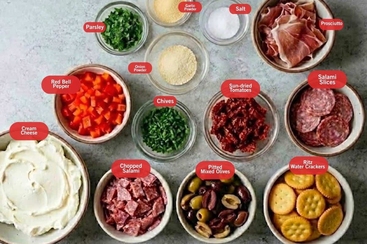 Flat lay photo of ingredient shot of parsley, onion and garlic powder, salt, prosciutto, salami slices, ritz water crackers, pitted mixed olives, chopped salami, cream cheese, red bell pepper, chives, chopped sun-dried tomatoes - 2