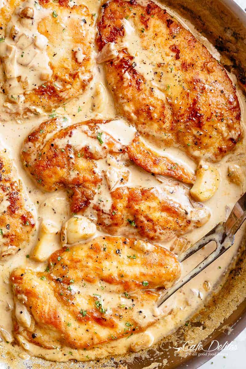 A silver frying pan of deliciously five Chicken Breasts in a garlic cream sauce with whole garlic cloves and a silver serving fork. | cafedelites.com