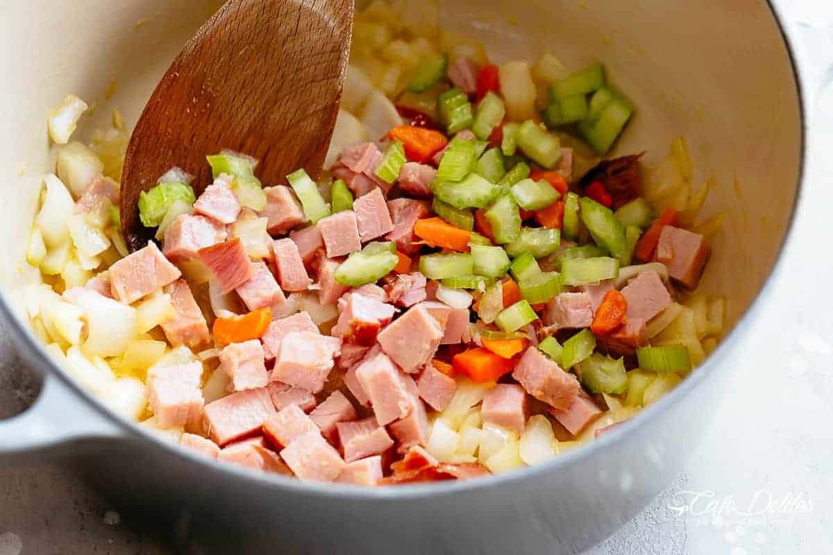 Chopped ham, celery, onion and garlic frying in a large pot, being stirred by a wooden spoon. 