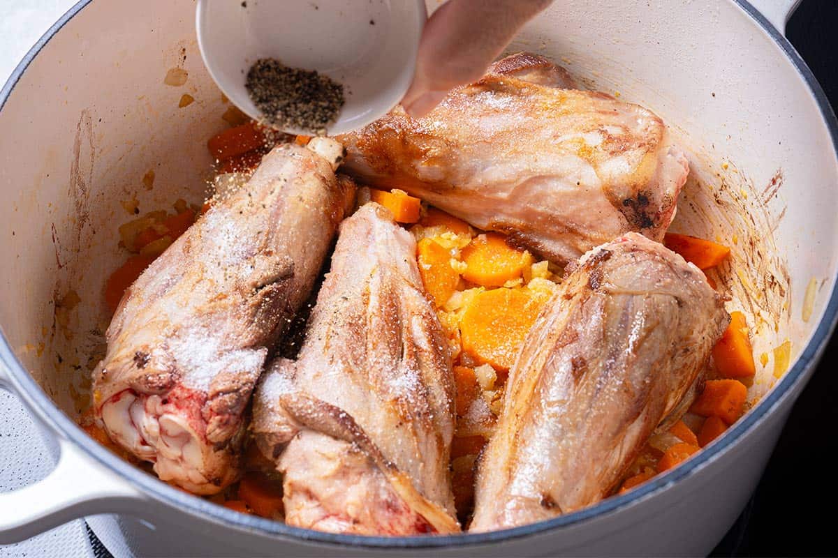 carrots, diced onion  and lamb shanks in a blue pot being sprinkled with salt and pepper