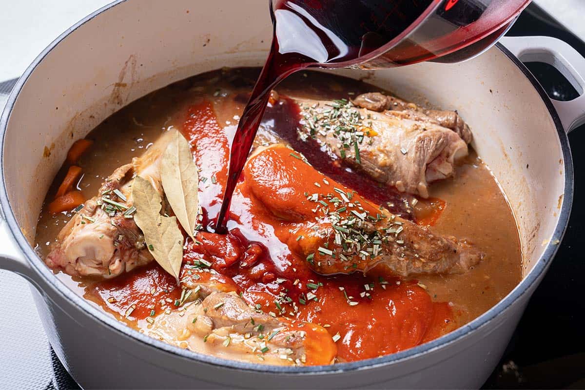 carrots, diced onion  and lamb shanks and stock in a blue pot with wine being poured in