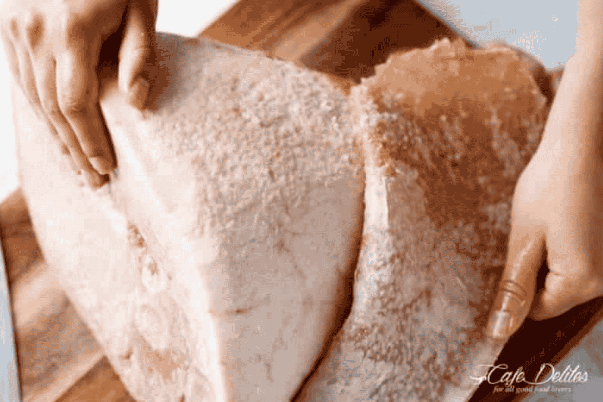 top down view of a leg of ham with bone in sitting on a board with a hand peeling off the layer of rind revealing a white fatty layer covering the leg