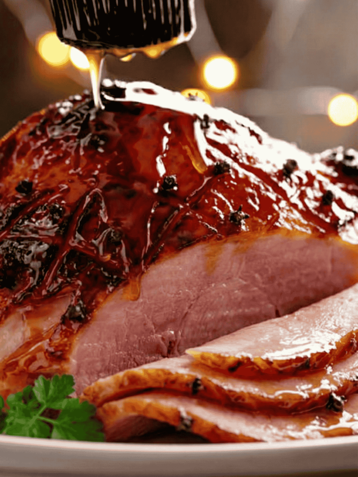 a side shot of a leg ham with slices cut out and a black brush covering it with a thick sticky clear glaze, the ham looks baked with black charred pieces speckled all over