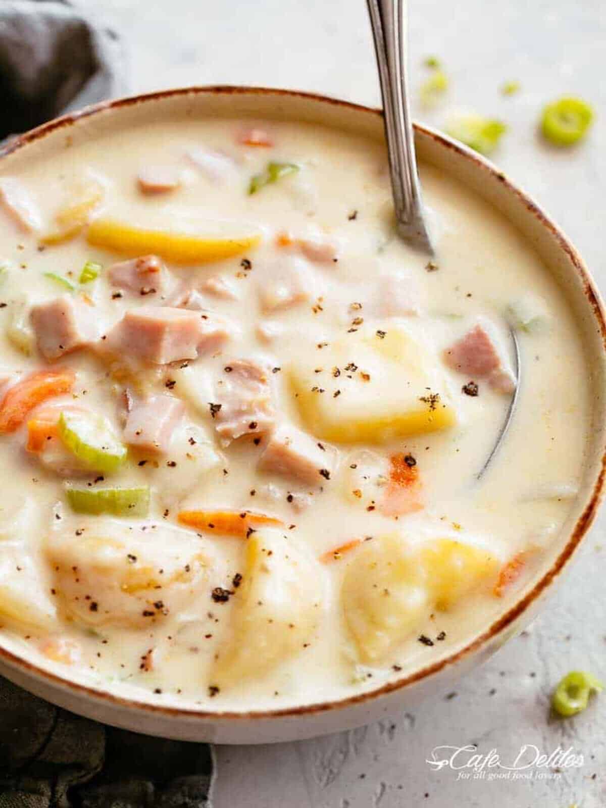 Image of a scrumptious creamy ham and potato soup with cracked pepper sprinkled on top, being stirred by a spoon.