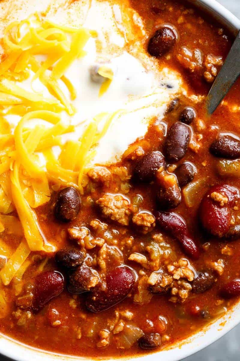 Ladling beef chili into a bowl topped with shredded cheese, cilantro, jalapeños, and lime.