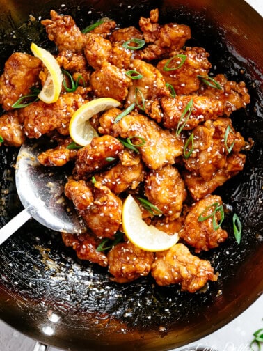 golden chicken bites cooked and covered in a sticky sauce with sesame seeds, sliced lemon wedges and chopped spring onion sprinkled on top. The chicken is being pushed around a wok with a metal spatula.