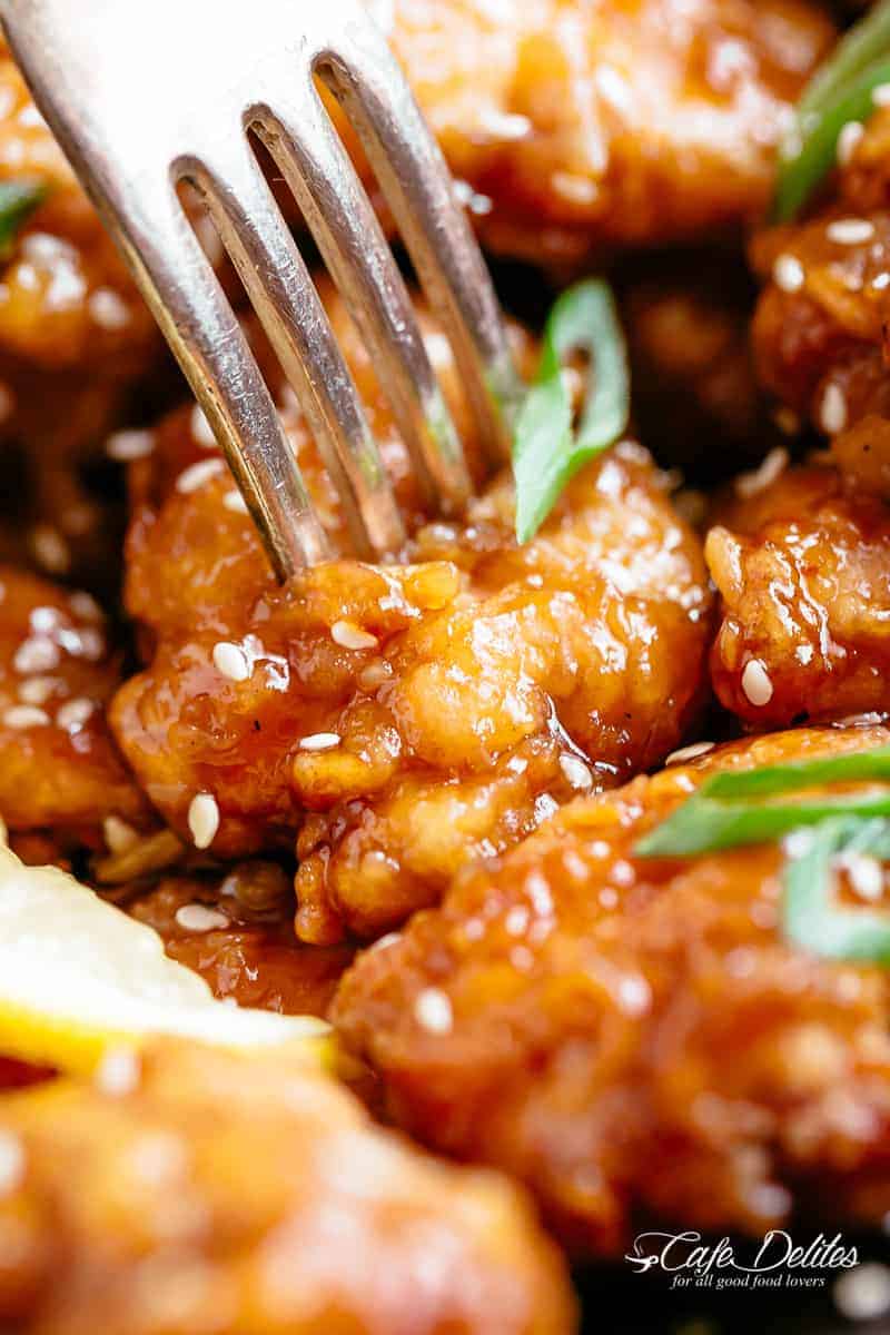 Close up of fork penetrating a crispy lemon chicken bite. The chicken is covered in sesame seeds and sliced spring onion. 