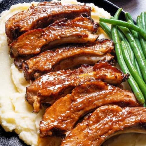 Slow-cooked barbecue ribs resting on a bed of mashed potatoes, accompanied by a side of green beans, served on a black serving plate.