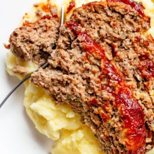 Top view image of A fork slicing through a piece of meatloaf and mashed potato on a white plate.