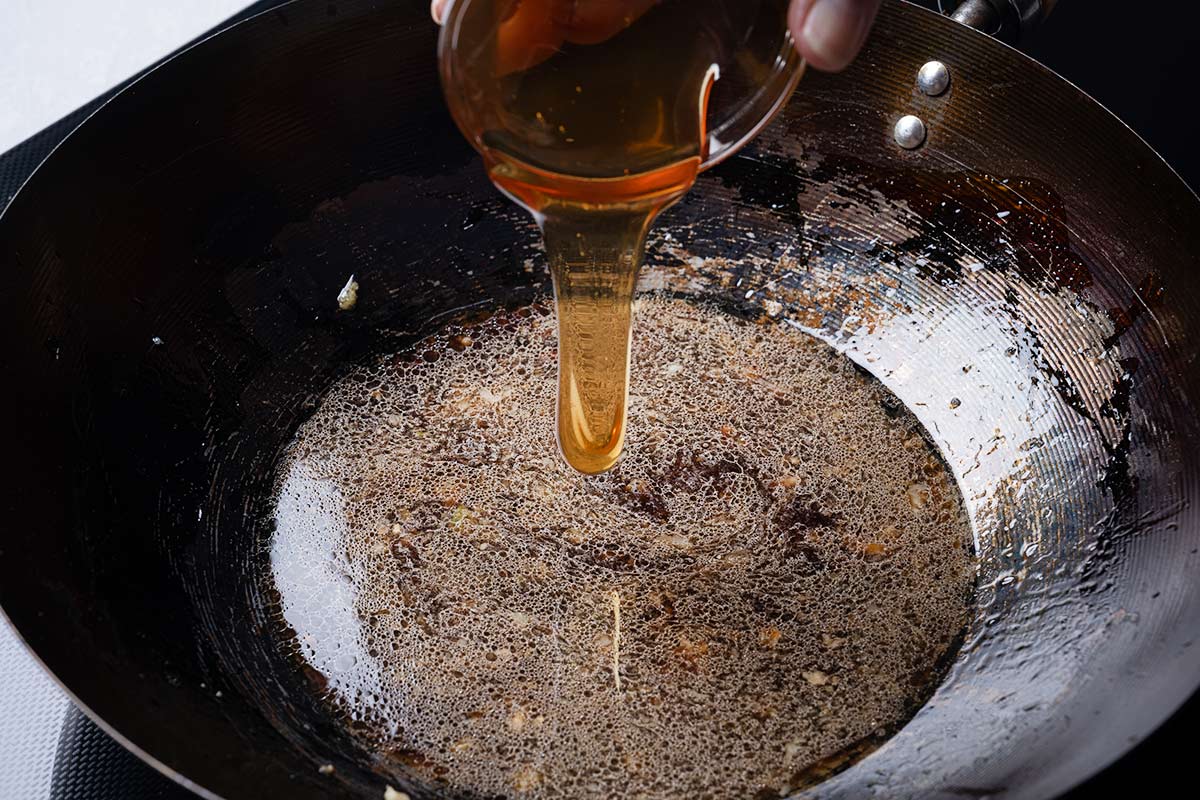 A wok with chicken Stock or broth, soy sauce, lemon juice, sugar and some salt with honey being poured in
