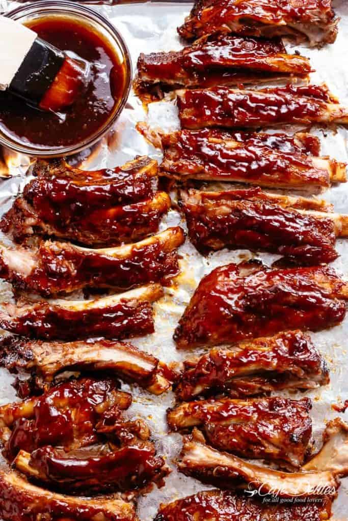 Slow cooker ribs cooked and separated on baking paper next to a ramekin of BBQ sauce with a basting brush dipped in it. 