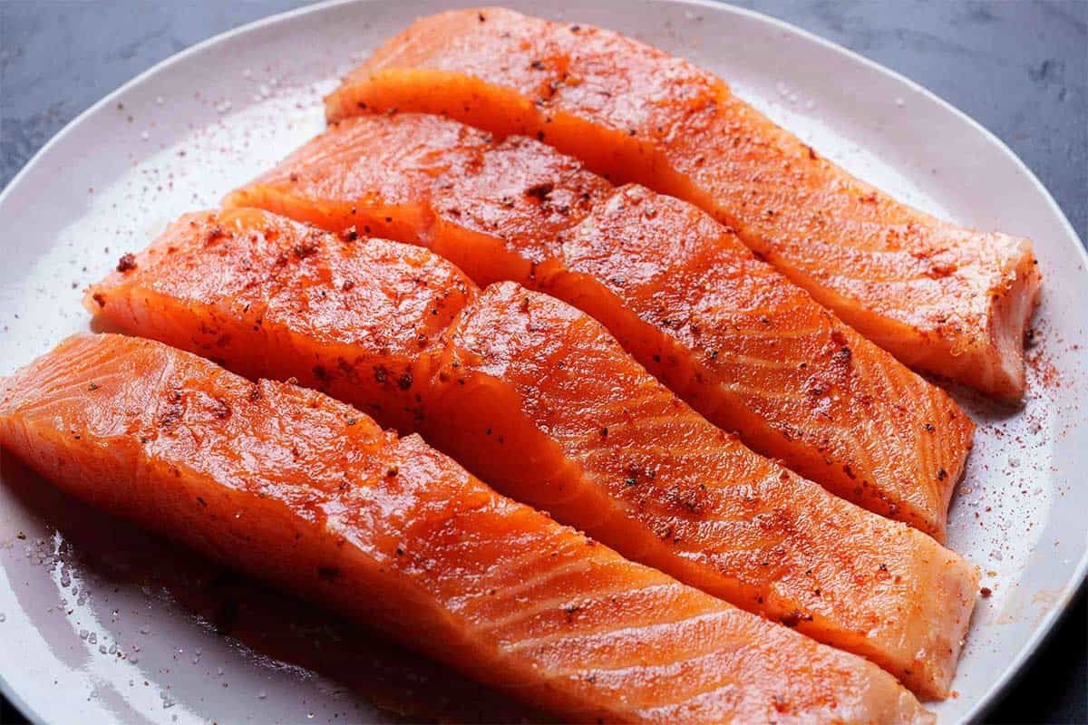 Image of beautifully laid-out salmon fillets being seasoned with salt, pepper, and paprika on a white circular plate.