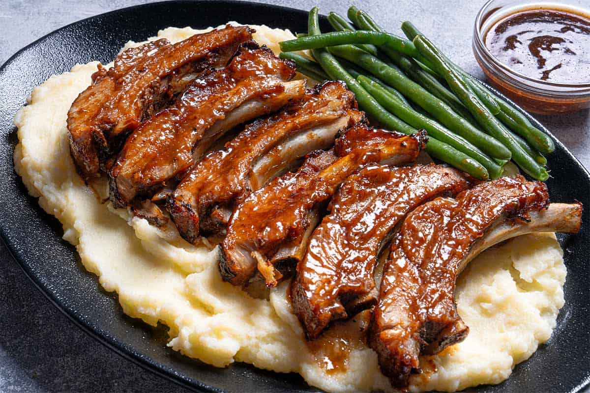 barbecue slow cooker ribs cut individually placed on mashed potato with beans on the side and barbecue sauce nexto the plate