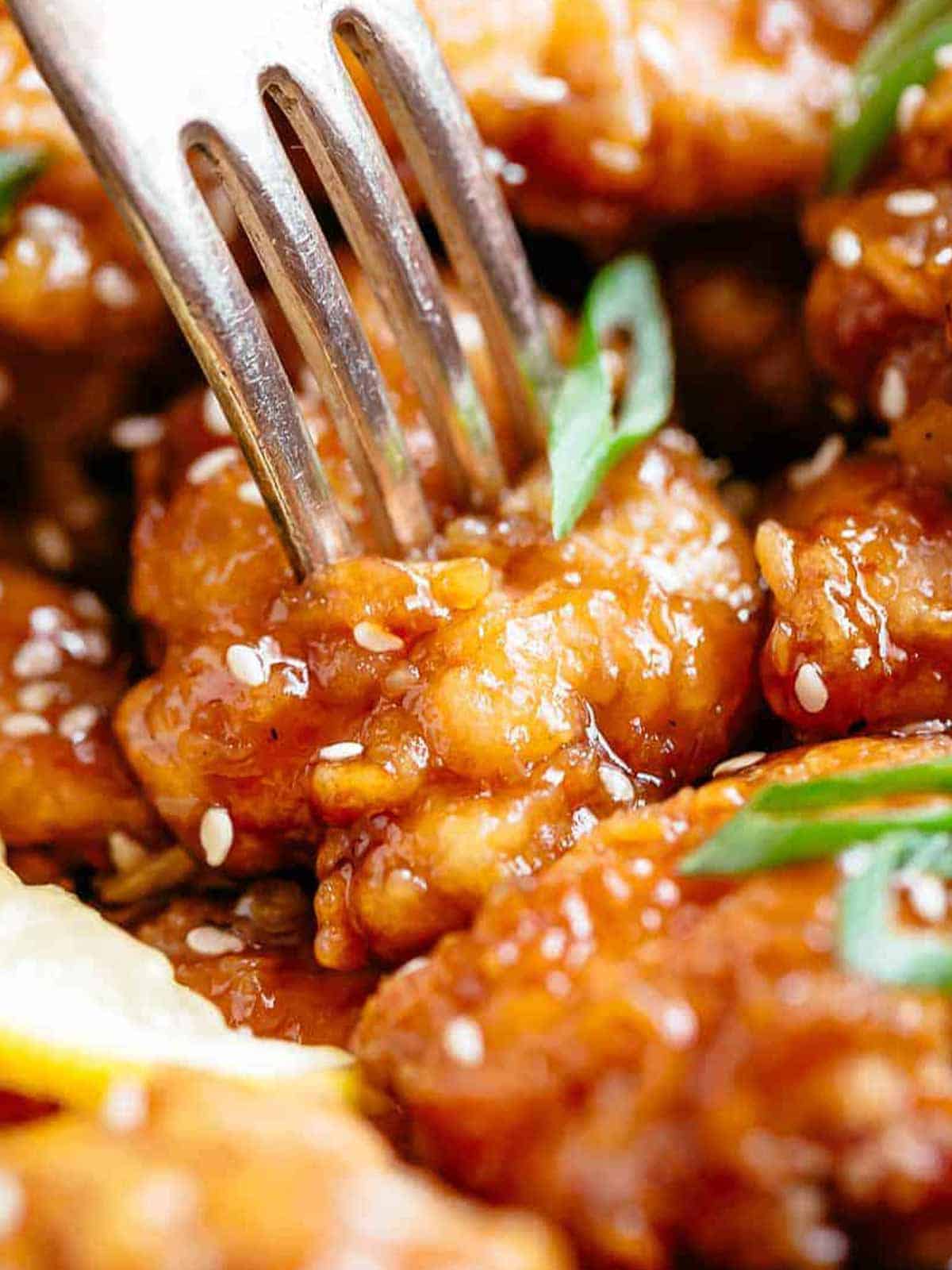 Close up of fork penetrating a crispy lemon chicken bite. The chicken is covered in sesame seeds and sliced spring onion. Looks delicious