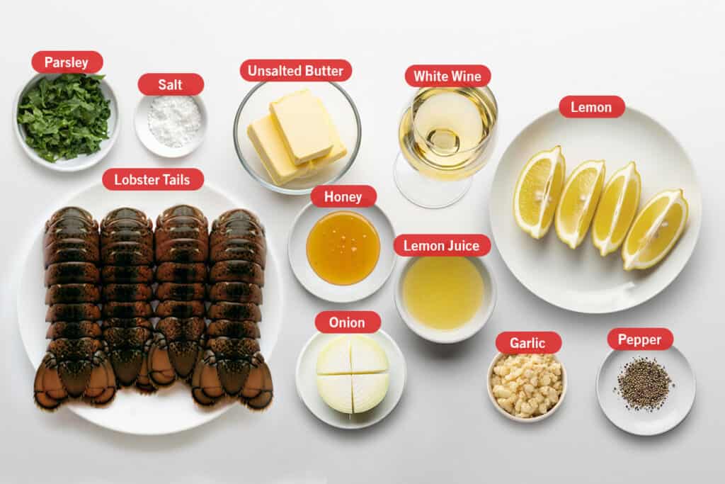 Flatlay of ingredients for broiled lobster tails, each in a separate bowl or plate. Includes six raw lobster tails on a white plate, a small bowl of chopped parsley, a bowl of salt, a dish of unsalted butter, a glass of white wine, lemon wedges on a plate, a small bowl of honey, a bowl of lemon juice, a quartered white onion, a small bowl of minced garlic, and a small plate of cracked black pepper. 
