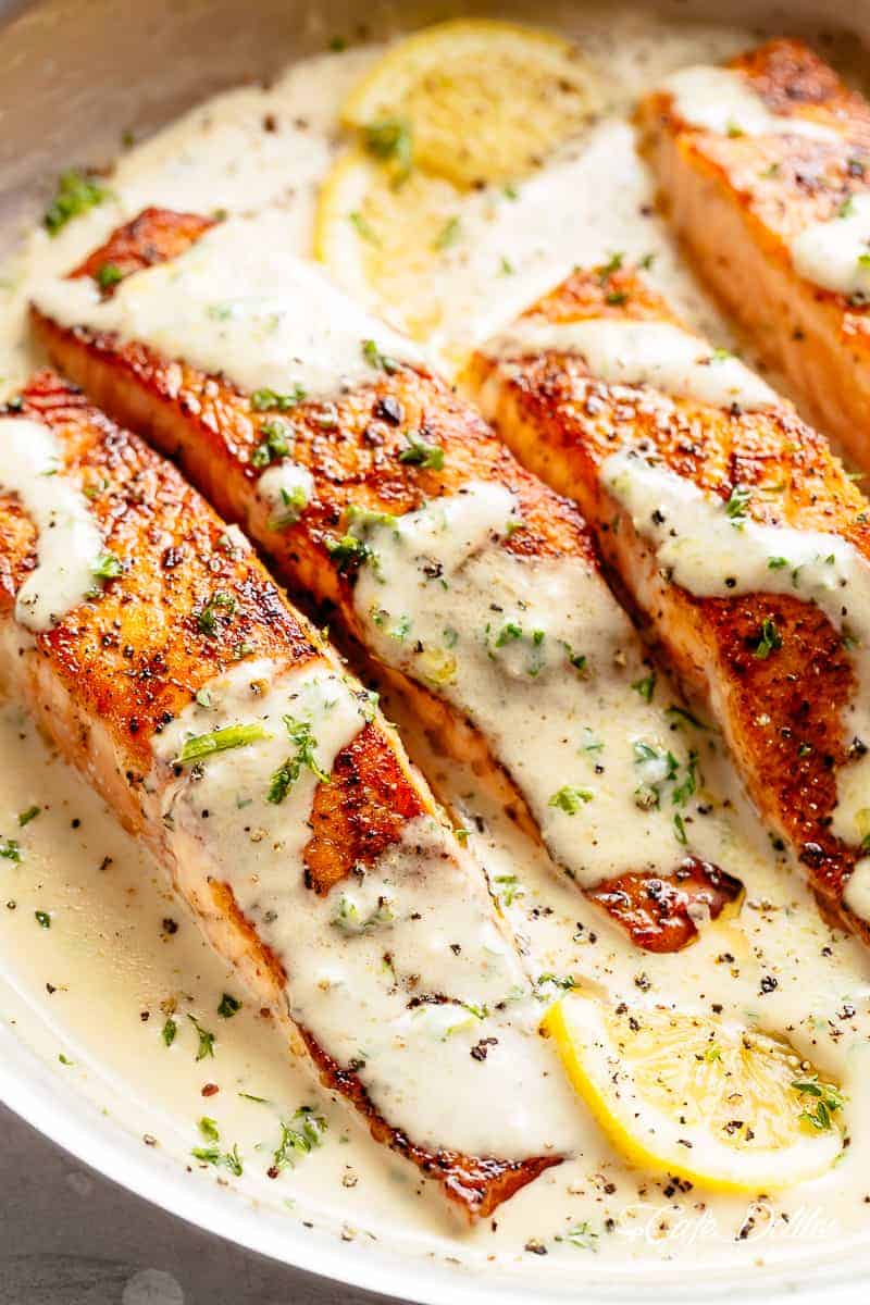 Four salmon fillets sitting and covered in creamy garlic butter sauce. Fresh herbs and two lemon wedges are scattered across the top of the fish. 