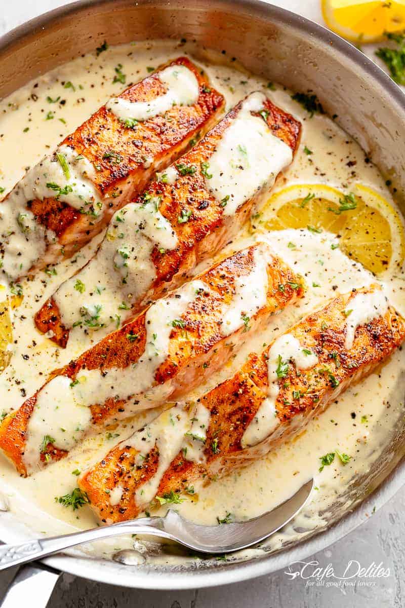 Four salmon fillets in a creamy garlic butter sauce in a large pan with a serving spoon resting inside the pan. Fresh green herbs are sprinkled over top of the fish and two lemon wedges sit beneath two of the fillets. 