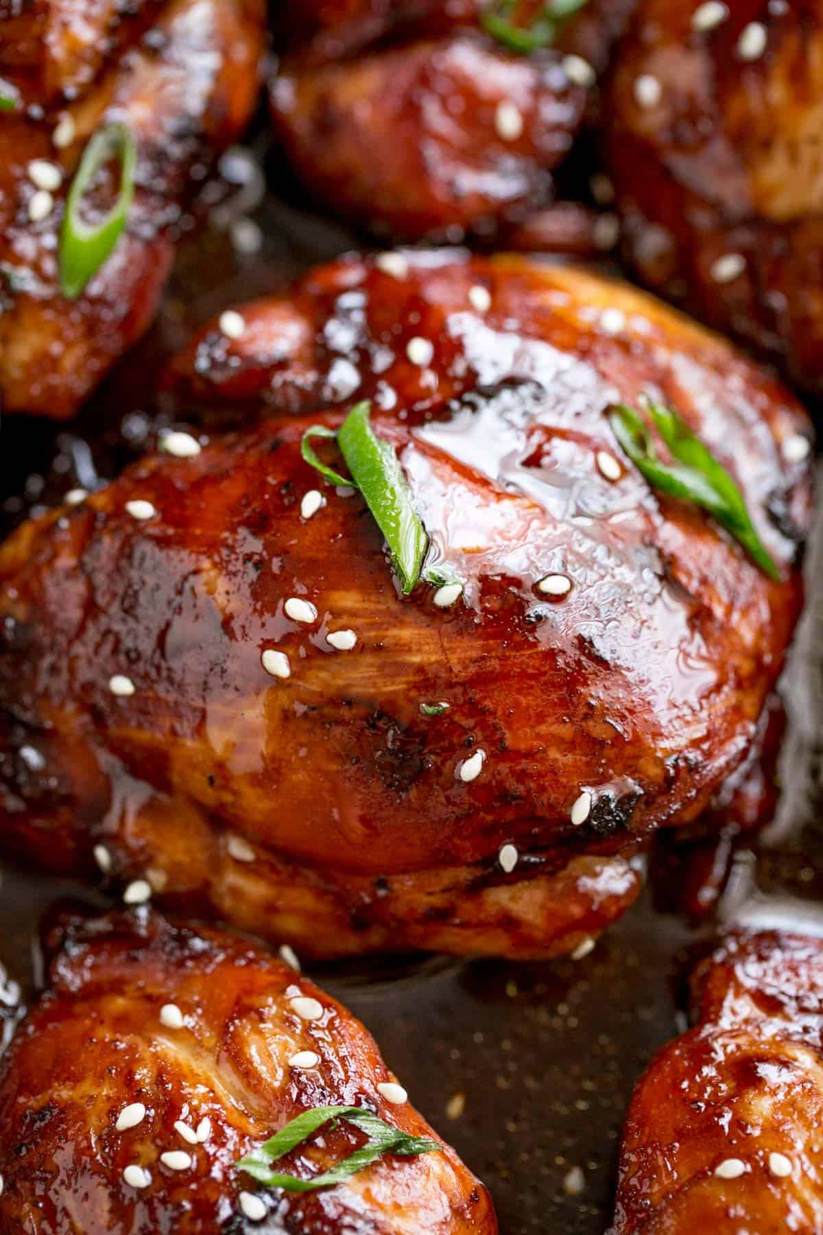 A close-up image of an oven-baked chicken thigh glazed with honey soy sauce and garnished with sesame seeds and scallions.