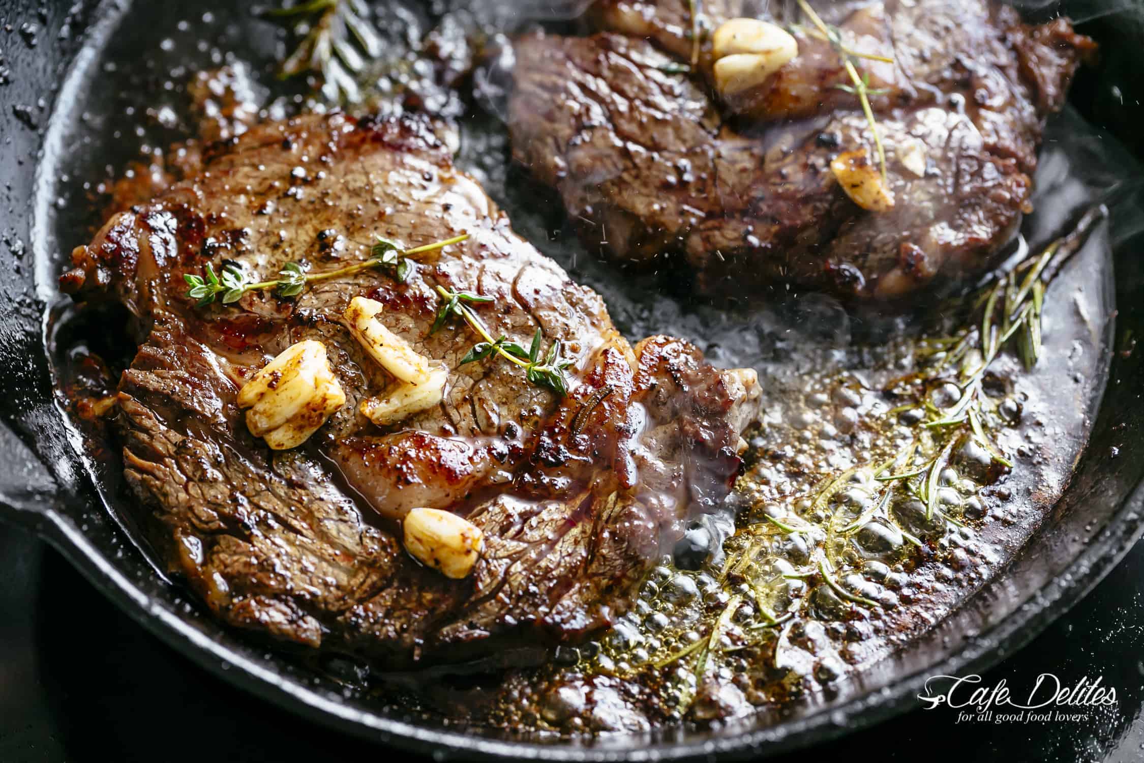 A close up shot of sizzling steaks covered in brown butter and garlic with a stick of rosemary simmering in a skillet.