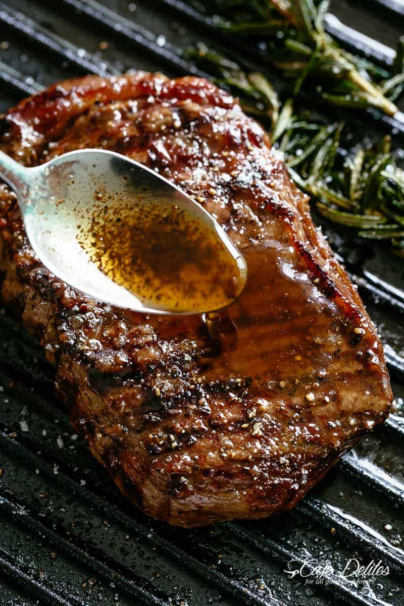 Juicy steak on a black grill with rosemary, with silver spoon drizzling garlic browned butter over steak. 