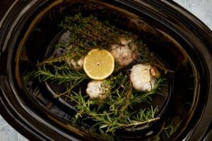 Garlic halves, thyme, rosemary, and lemon arranged in slow cooker base