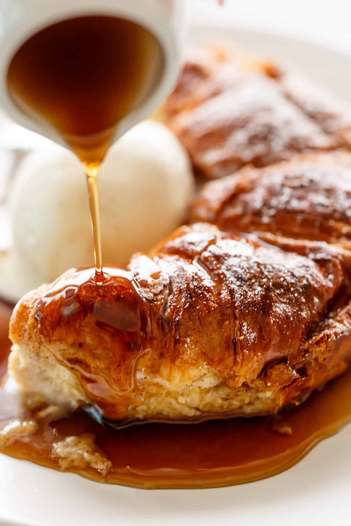 An image of a baked croissant, which has recently been taken out of the oven, is served on a white plate alongside a scoop of ice cream drizzled with vanilla sauce.