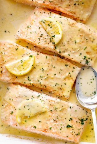 Four pieces of salmon covered in a creamy lemon sauce and topped with fresh parsley and slices of lemon. The salmon is arranged in a white baking dish, and a silver serving spoon is visible on the right side, partially submerged in the sauce. The salmon appears to be cooked to perfection, with a slightly browned and crispy top. The sauce is a pale yellow color and appears rich and creamy.