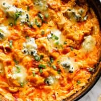 This close-up shot shows a cast iron skillet filled with a creamy, cheesy buffalo chicken dip. The dip is a vibrant orange color, with melted cheese, chunks of chicken, and green onion scallions sprinkled throughout. There are also generous dollops of blue cheese sprinkled on top. The edges of the skillet are darkened, suggesting the dip has been baked. The background is a marble surface, a few pieces of celery, and the corner of a cracker. Everything looks yummy