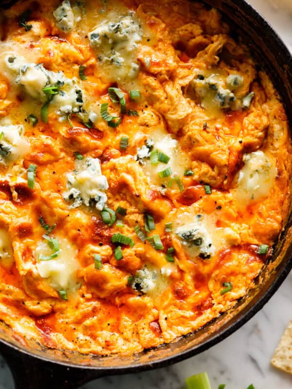 This close-up shot shows a cast iron skillet filled with a creamy, cheesy buffalo chicken dip. The dip is a vibrant orange color, with melted cheese, chunks of chicken, and green onion scallions sprinkled throughout. There are also generous dollops of blue cheese sprinkled on top. The edges of the skillet are darkened, suggesting the dip has been baked. The background is a marble surface, a few pieces of celery, and the corner of a cracker. Everything looks yummy
