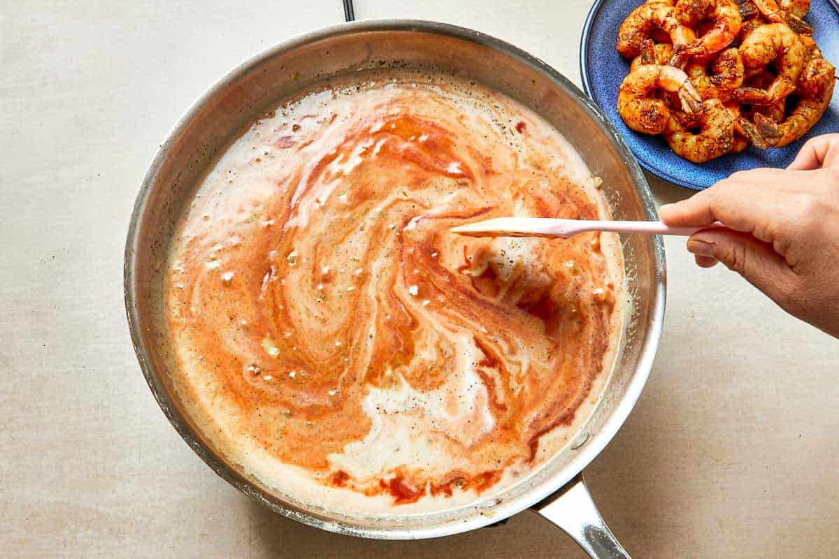 Hand stirs shrimp in a pan with a thickening sauce of crushed tomatoes, coconut milk, and chili powder.