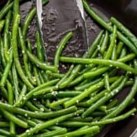 Sautéed green beans with a sprinkle of course salt in a pan being pushed aside by a pair of tongs.