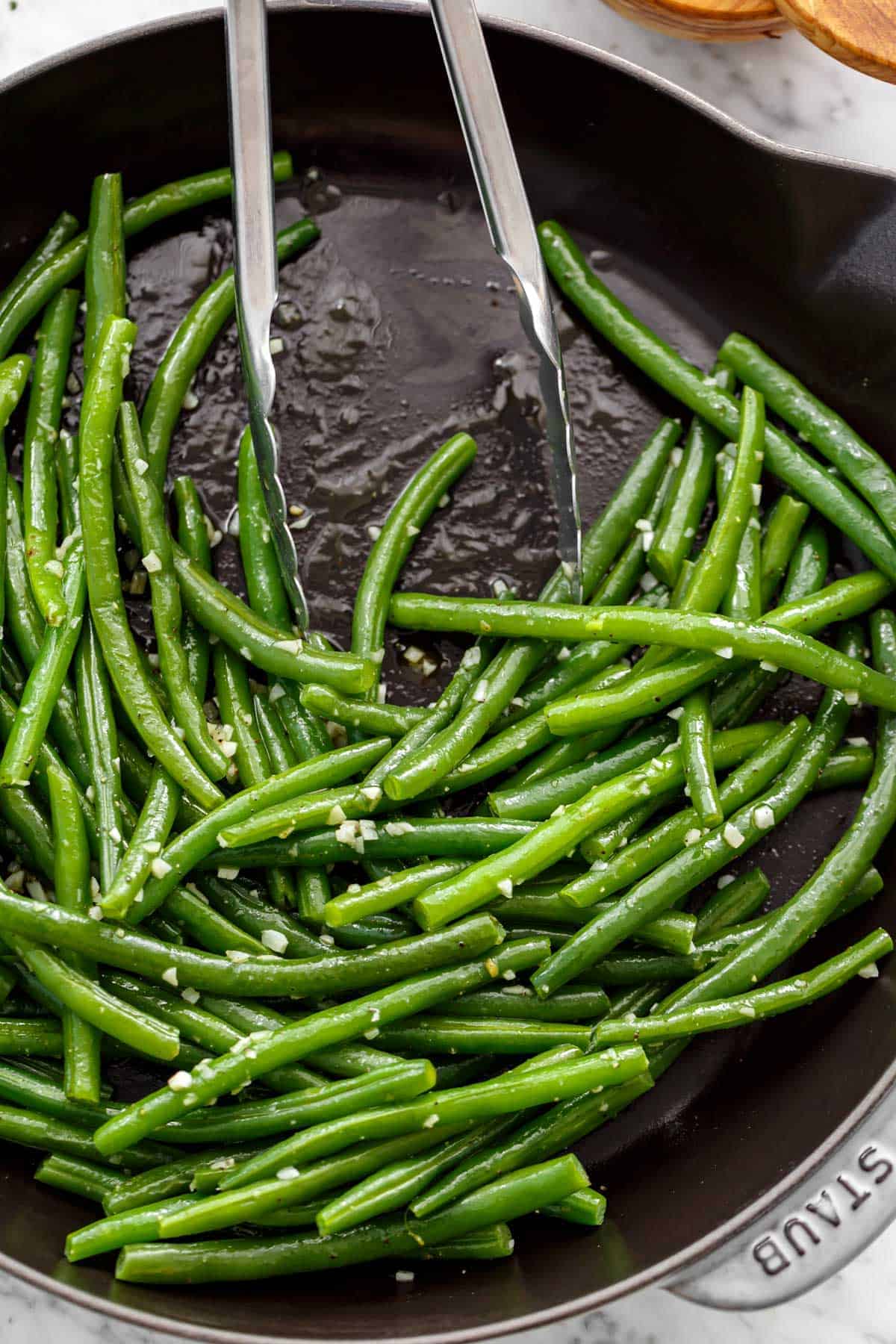 Pan of sautéed, buttery green beans covered in garlic with tongs leaning on side of pan.