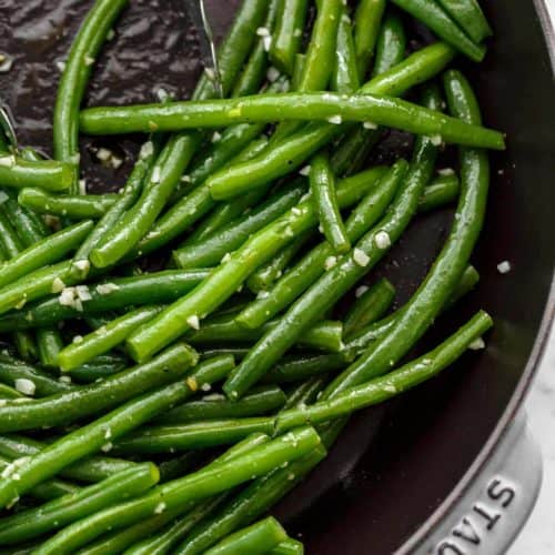 Close up image of Sautéed green beans with a sprinkle of course salt in a pan being pushed aside by a pair of tongs.