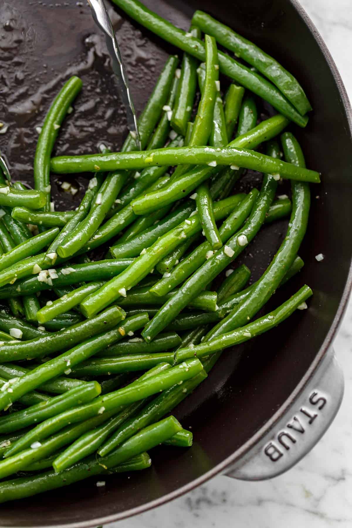 Close up of Sautéed green beans with a sprinkle of course salt in a pan being pushed aside by a pair of tongs.