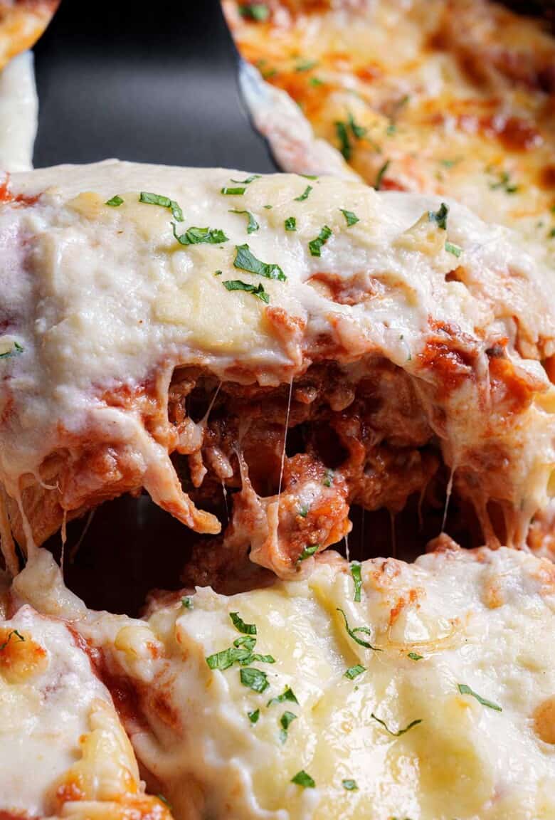 A beautiful close-up of a slice of lasagna being lifted by a spatula from the baking tray, showcasing layers of pasta, rich meat sauce, creamy cheese, and a golden, bubbly top.