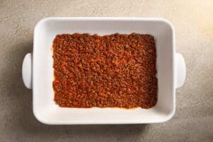 A white rectangular baking dish with a thin, even layer of rich red meat sauce spread across the bottom.
