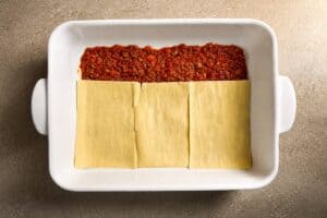 A white baking dish showing three flat, dry lasagna noodles placed side-by-side on top of a layer of meat sauce.