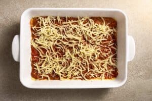 A close-up of a lasagna layer in a baking dish, featuring red meat sauce generously sprinkled with shredded white mozzarella cheese.