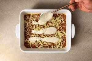 A baking dish filled with lasagna, showing the top layer of meat sauce being covered with thick swirls of creamy white sauce.
