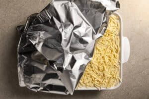 A white baking dish filled with unbaked lasagna, topped with an even layer of shredded cheese, being covered with aluminum foil.