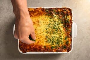 A freshly baked, golden-brown lasagna in a white dish, with a hand sprinkling chopped green parsley over the bubbly, melted cheese.