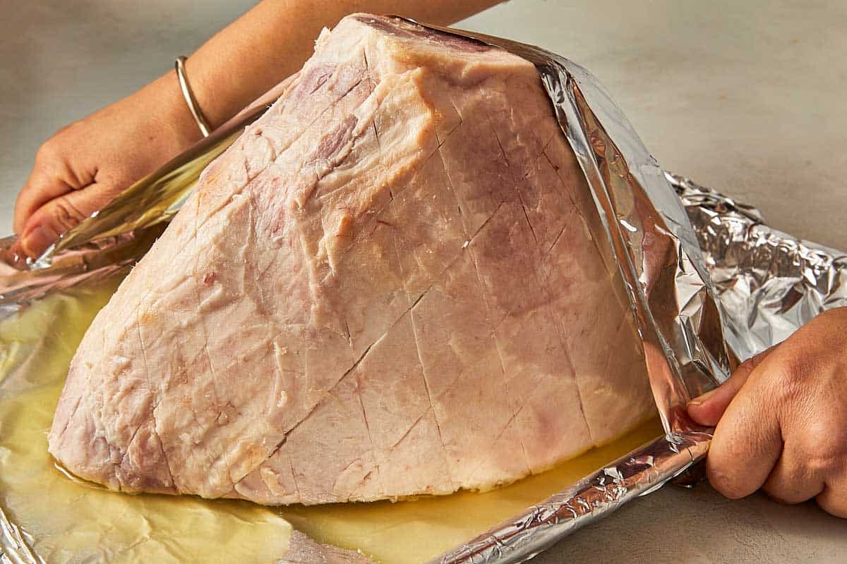 A person uses a knife to cut a large ham placed on foil, highlighting the preparation of the meat.